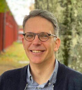 A smiling man in a dark jacket and shirt stands smiling by a tree.